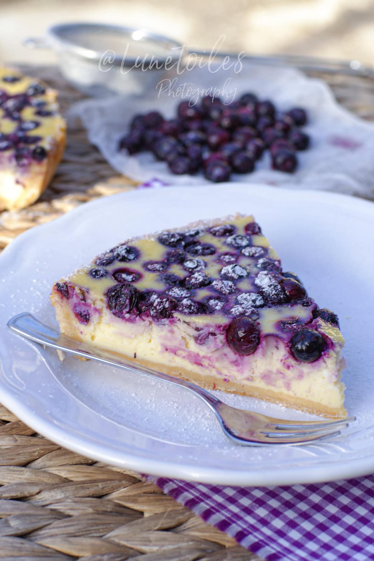 Part de cheesecake aux myrtilles marbré violet, saupoudrée de sucre glace, servie sur une assiette blanche avec une fourchette; en arrière-plan, des myrtilles sur un linge et un petit tamis, lumière naturelle.