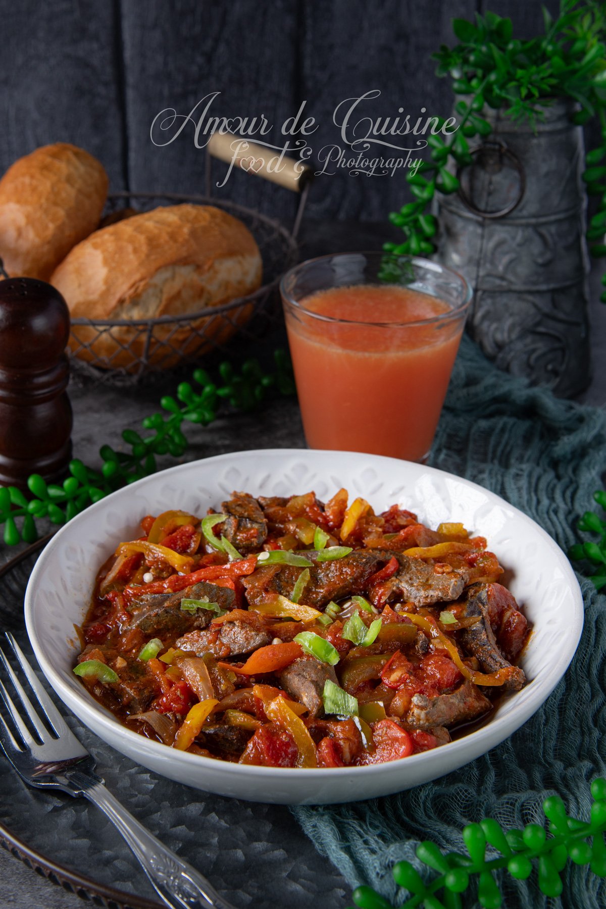 Assiette de kebda à la chakchouka : foie d’agneau sauté dans une sauce tomate aux poivrons et oignons, garni d’herbes, avec du pain en arrière-plan et un verre de jus.