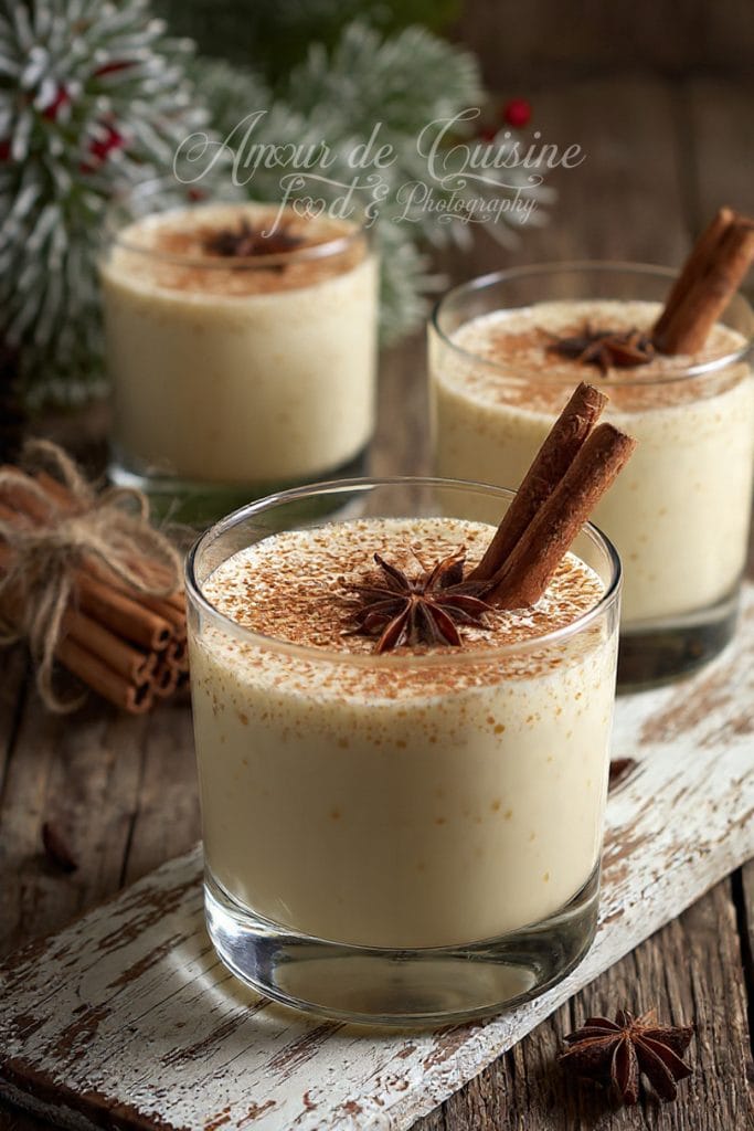 Verres de lait de poule crémeux garnis de bâtons de cannelle et d’anis étoilé, posés sur une planche en bois vieillie, avec en arrière-plan des décorations de sapin givré et des épices d’hiver pour une ambiance chaleureuse et festive.