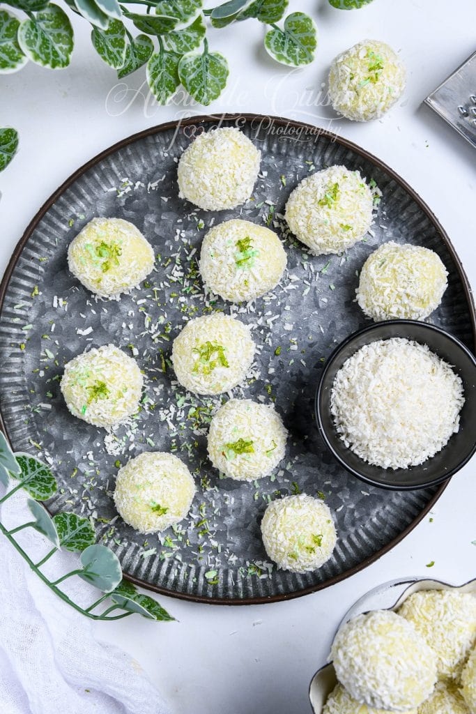 Overhead view of Key Lime Pie truffles coated in shredded coconut, arranged on a dark round plate with lime zest sprinkled on top. A small black bowl filled with coconut sits to the side, surrounded by fresh green leaves and a light white cloth.