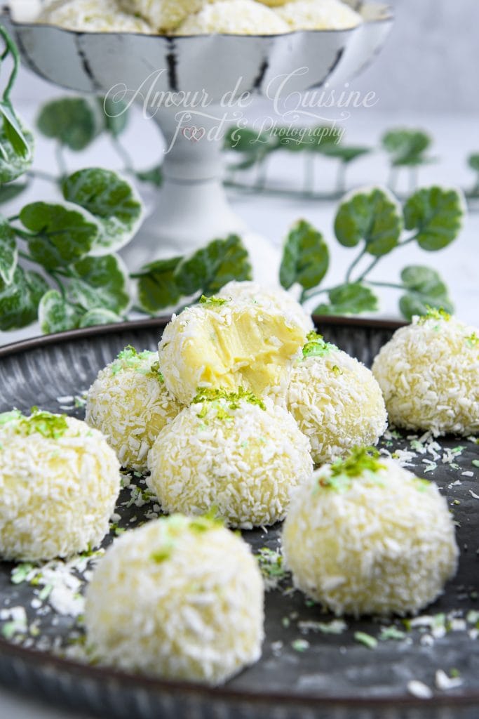 Close-up of round Key Lime Pie truffles coated in shredded coconut, arranged on a dark plate. One truffle is bitten open, showing its creamy lime-infused center. Fresh lime zest is sprinkled on top, with a blurred decorative plant and serving dish in the background.