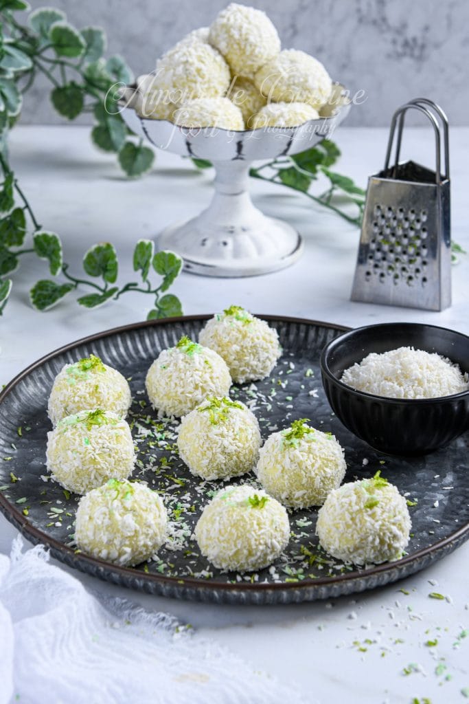 Display of Key Lime Pie truffles coated in shredded coconut, arranged on a dark serving plate with fresh lime zest sprinkled on top. A black bowl filled with coconut sits beside them, while more truffles are stacked in a white pedestal dish in the background. Decorative green leaves and a metal grater complete the bright, fresh kitchen scene.
