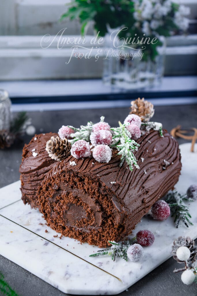 Bûche de Noël au chocolat roulée, recouverte de ganache striée façon écorce, décorée de canneberges et branches de romarin cristallisées, posée sur une planche en marbre avec décor hivernal.