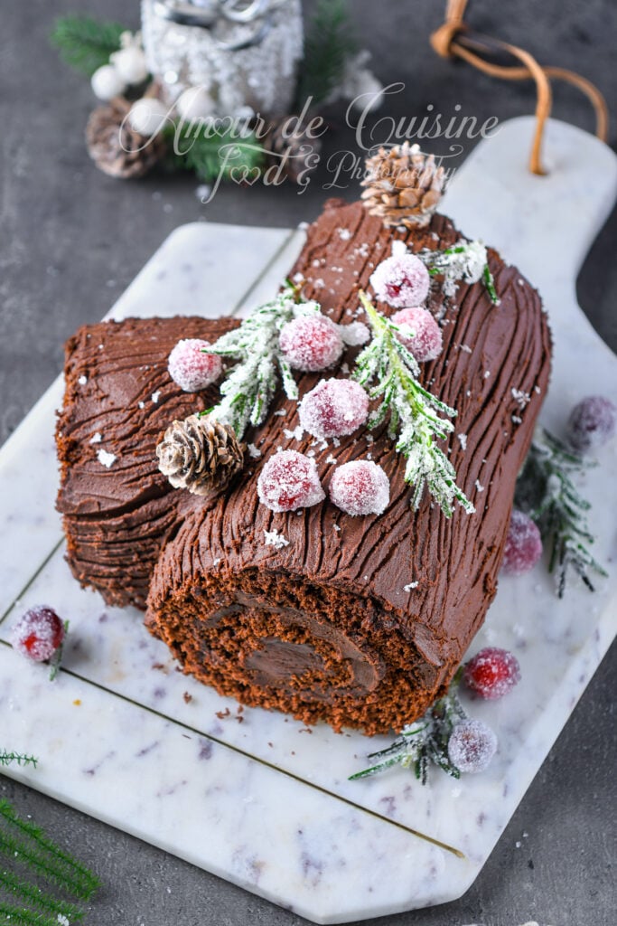 Bûche de Noël au chocolat vue de dessus, roulée et recouverte de ganache striée façon écorce, décorée de canneberges cristallisées, de brins de romarin givrés et d’une petite pomme de pin, posée sur une planche en marbre.