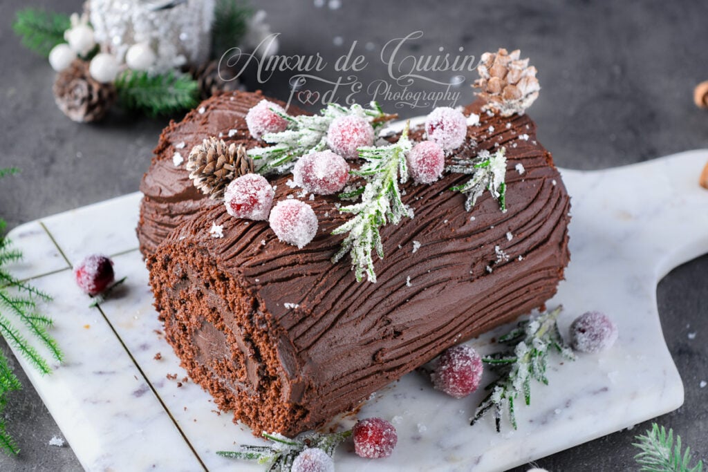 Bûche de Noël au chocolat posée sur une planche en marbre, recouverte de ganache striée façon écorce et décorée de canneberges cristallisées, de brins de romarin givrés et de petites pommes de pin pour un décor hivernal.