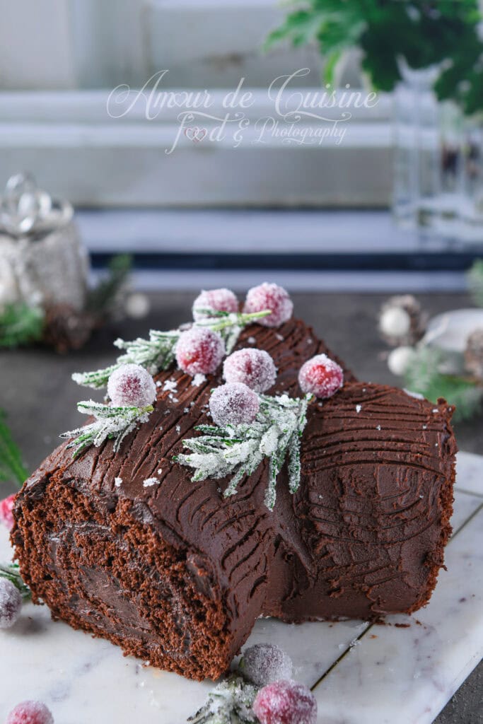 Bûche de Noël au chocolat vue de près, génoise roulée recouverte de ganache striée, garnie de canneberges rouges et de brins de romarin cristallisés, posée sur une planche en marbre avec décor hivernal flou en arrière-plan.