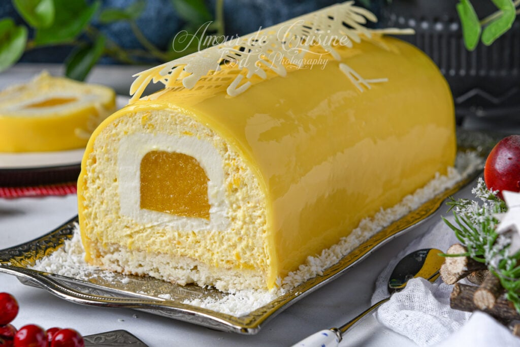 Bûche mangue coco entière au glaçage miroir jaune brillant, avec insert mangue visible à la coupe, mousse coco et base dacquoise, décorée d’un motif en chocolat blanc et posée sur un plat entouré de noix de coco râpée.