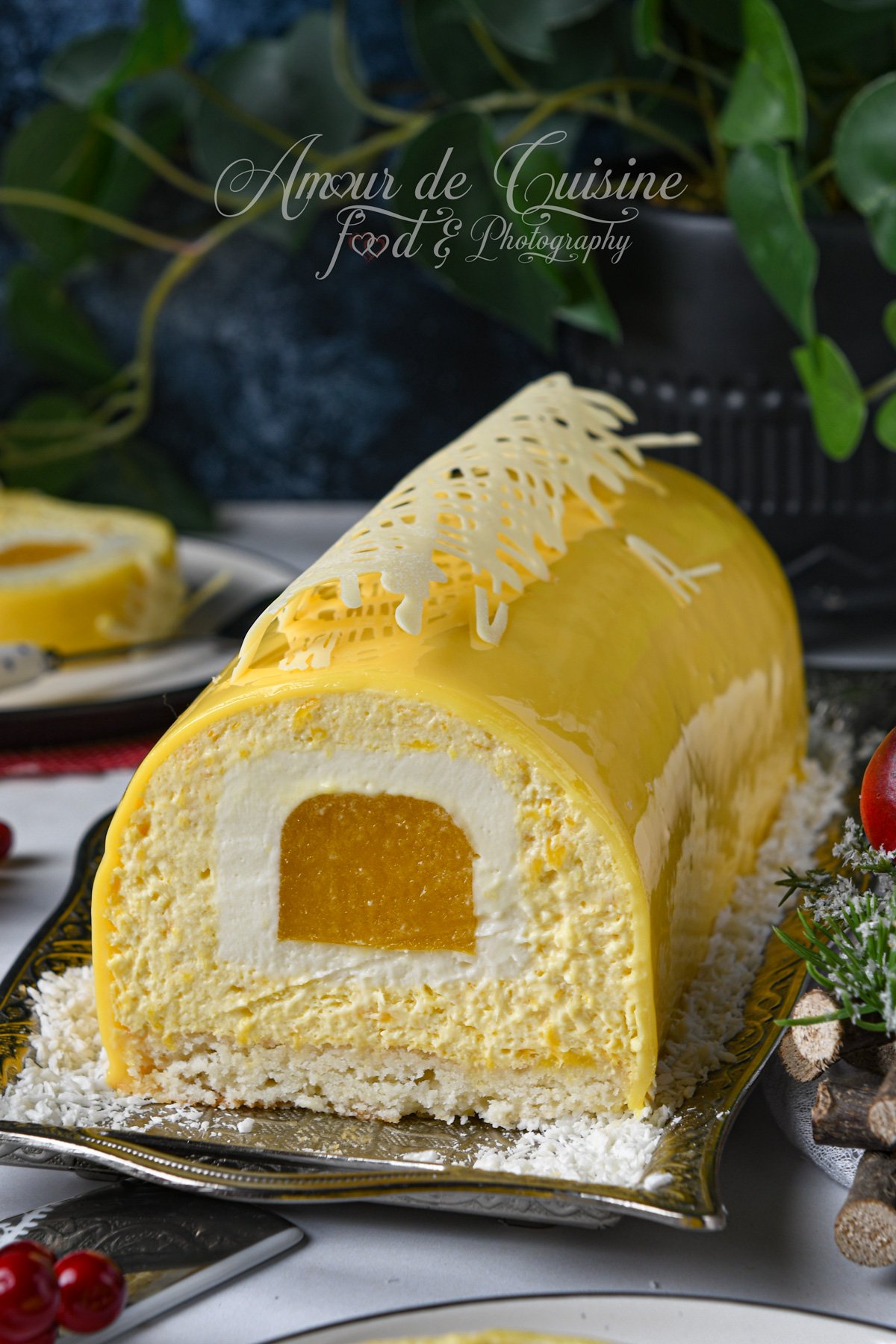 Bûche mangue coco au glaçage miroir jaune, coupée pour montrer l’insert mangue au centre, entourée de mousse coco et posée sur une base dacquoise, décorée d’une dentelle en chocolat blanc sur un plat avec noix de coco râpée.