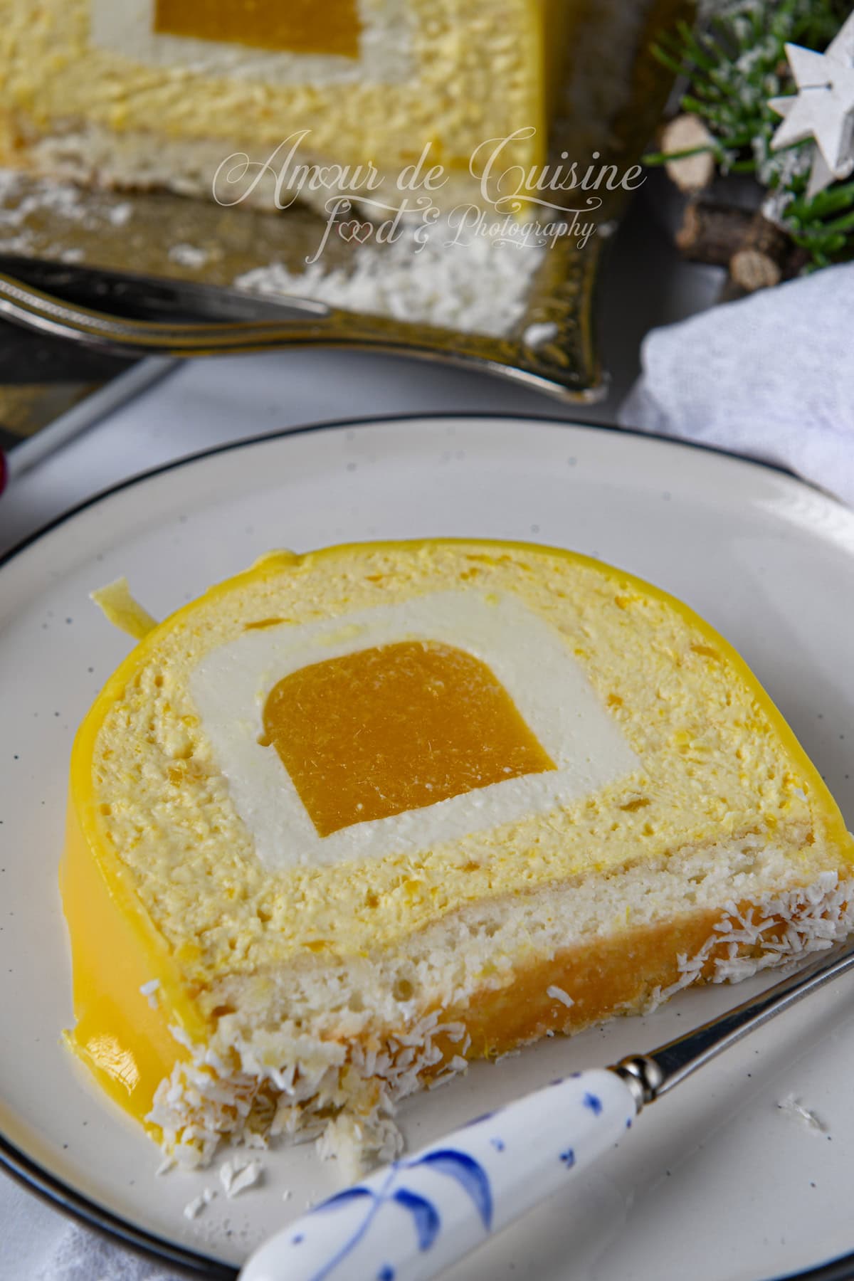 Tranche de bûche mangue coco au glaçage miroir jaune, avec insert mangue au centre, mousse coco et base dacquoise, servie dans une assiette avec un peu de noix de coco râpée.