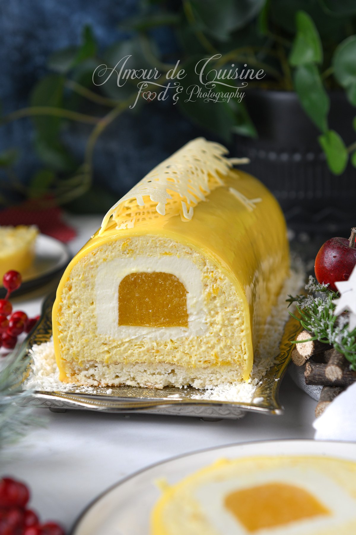 Part de bûche mangue coco au glaçage miroir jaune, avec insert mangue au centre, mousse coco, dacquoise coco en base et décor en chocolat blanc, présentée sur un plat avec noix de coco râpée.