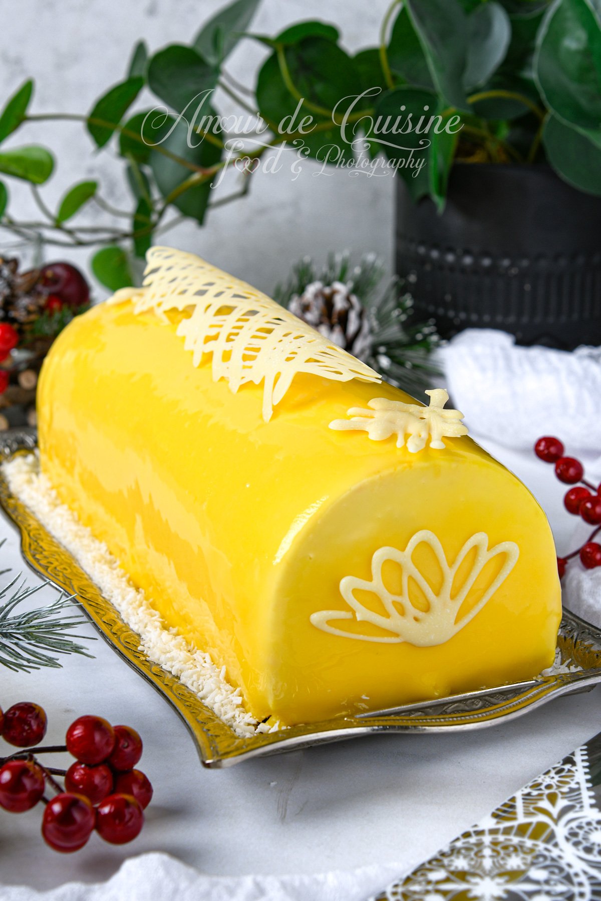 Bûche mangue coco au glaçage miroir jaune brillant, décorée de dentelle en chocolat blanc sur le dessus, posée sur un plat argenté avec un lit de noix de coco râpée et une mise en scène festive (baies rouges, feuillage).
