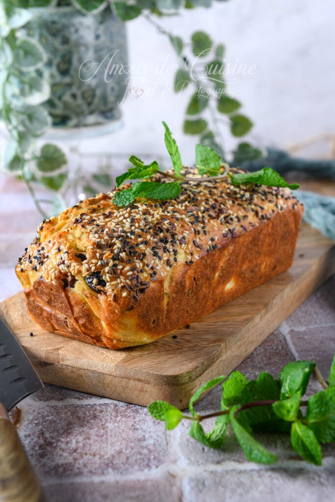 Cake salé à la feta, menthe et olives Kalamata entier, bien doré et recouvert de graines variées, décoré de feuilles de menthe fraîche, posé sur une planche en bois avec un fond végétal flou.