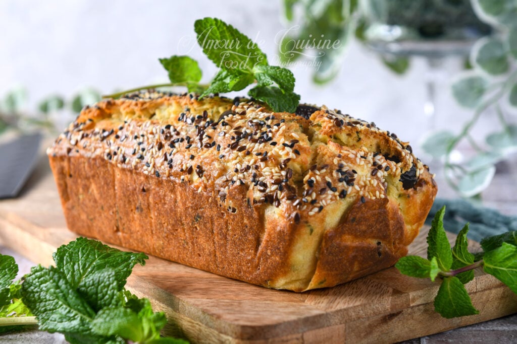 Cake salé à la feta, menthe et olives Kalamata entier, bien doré et gonflé, recouvert de graines variées et décoré de menthe fraîche, posé sur une planche en bois avec des herbes en avant-plan.