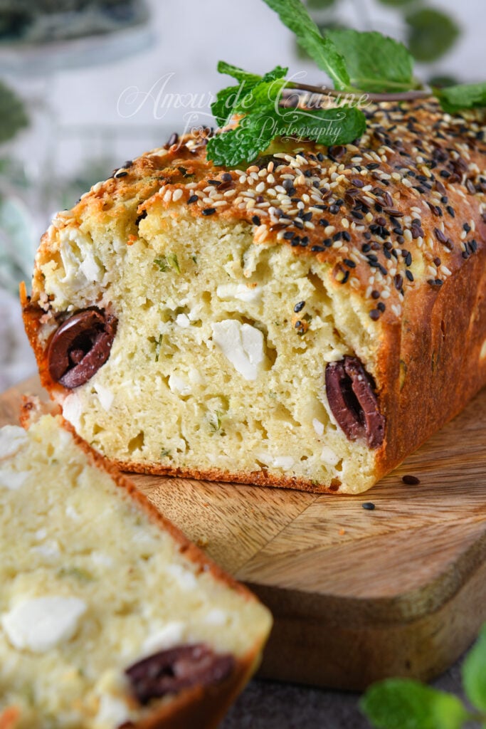 Tranche de cake salé à la feta, menthe et olives Kalamata, moelleux et parsemé de graines variées sur le dessus, posé sur une planche en bois avec une branche de menthe fraîche en décoration.