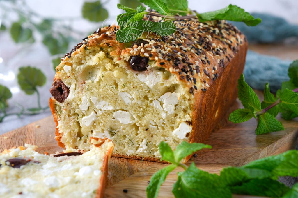 Gros plan sur un cake salé à la feta, menthe et olives Kalamata, coupé pour montrer une mie moelleuse garnie de dés de feta, recouvert de graines variées, avec une tranche au premier plan et des feuilles de menthe autour.