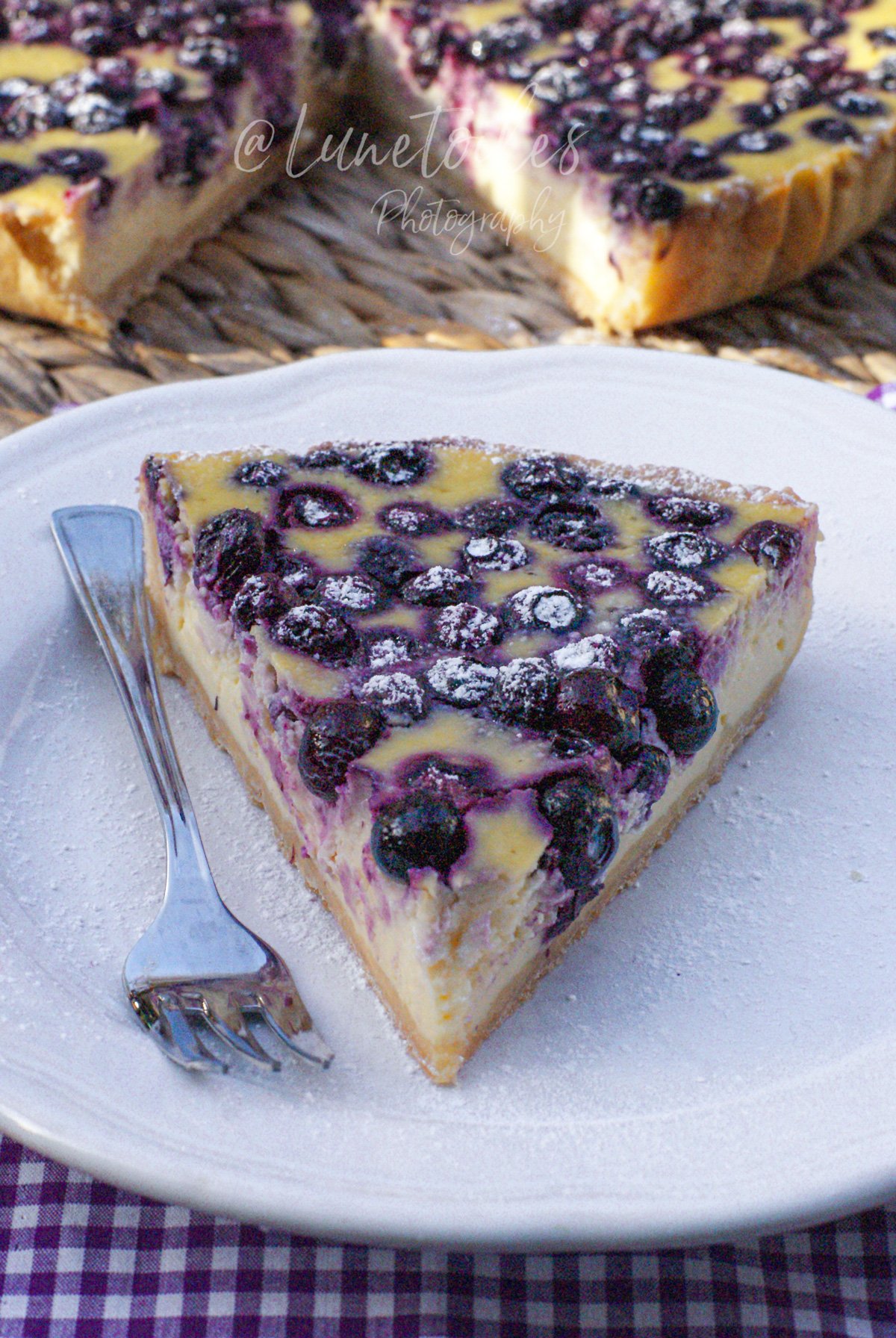 Part de cheesecake aux myrtilles saupoudrée de sucre glace, posée sur une assiette blanche avec une fourchette; en arrière-plan, le cheesecake entamé sur une table en fibres tressées, avec une nappe vichy violette.