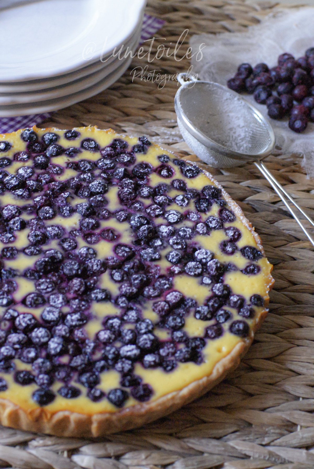 Cheesecake entier aux myrtilles, surface couverte de fruits, sur une table en fibres tressées; à côté, un petit tamis et des myrtilles posées sur un linge, avec des assiettes empilées en arrière-plan.