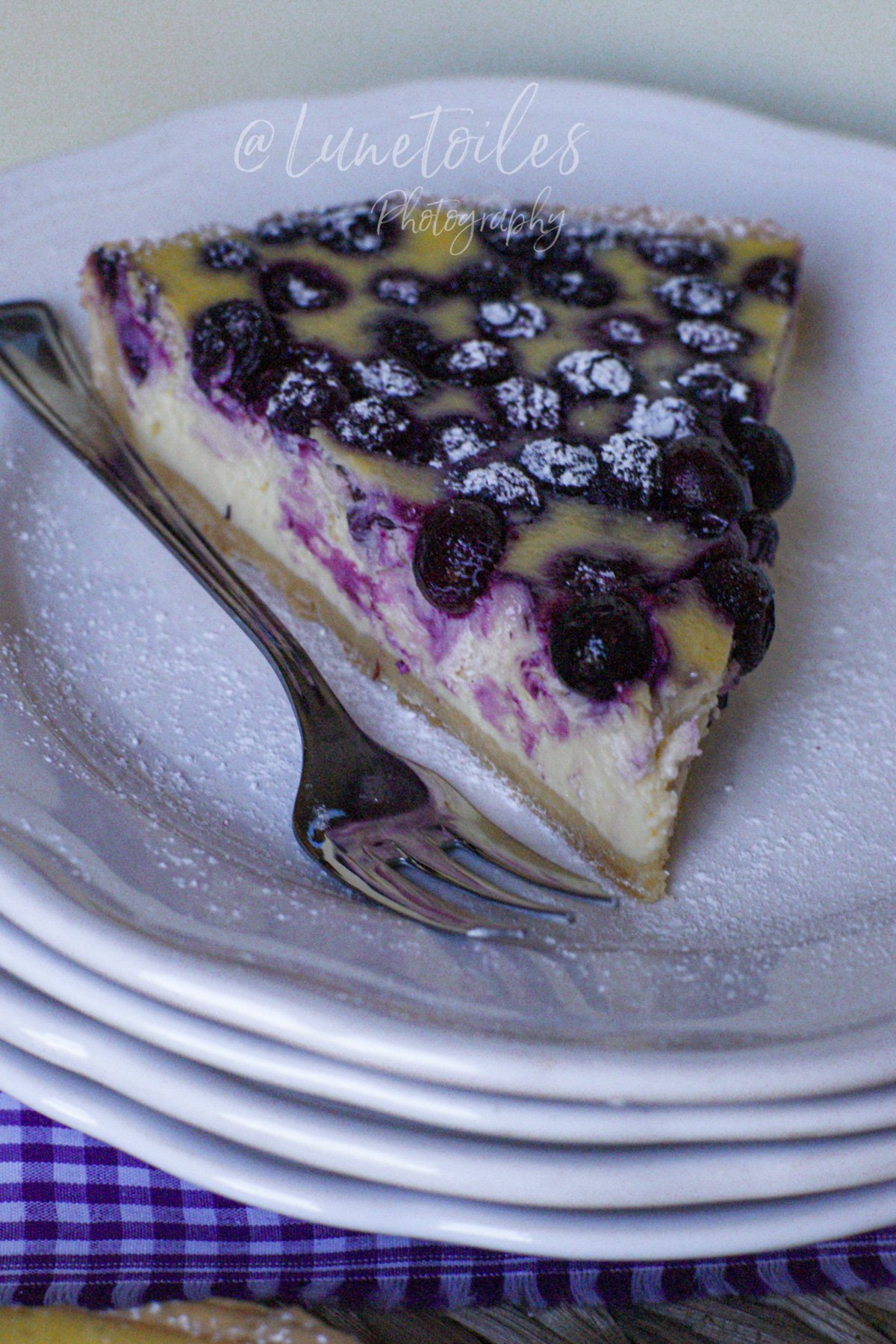Part de cheesecake aux myrtilles, marbrée de violet, posée sur une assiette blanche saupoudrée de sucre glace, avec une fourchette glissée dessous; assiettes empilées et nappe vichy violette en bas de l’image.