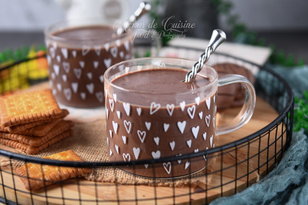 Deux tasses en verre décorées de cœurs, remplies de chocolat chaud parisien épais, avec une cuillère torsadée, servies sur un plateau avec biscuits et une douceur au chocolat.