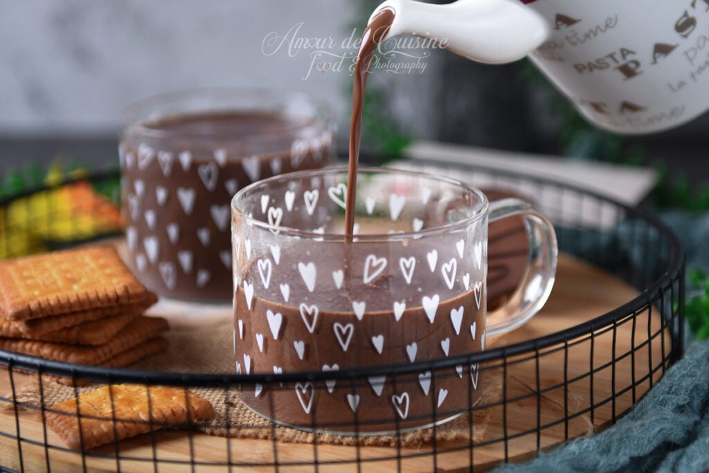 Un chocolat chaud parisien épais est versé d’une petite théière dans une tasse en verre décorée de cœurs, sur un plateau avec biscuits, avec une seconde tasse en arrière-plan.