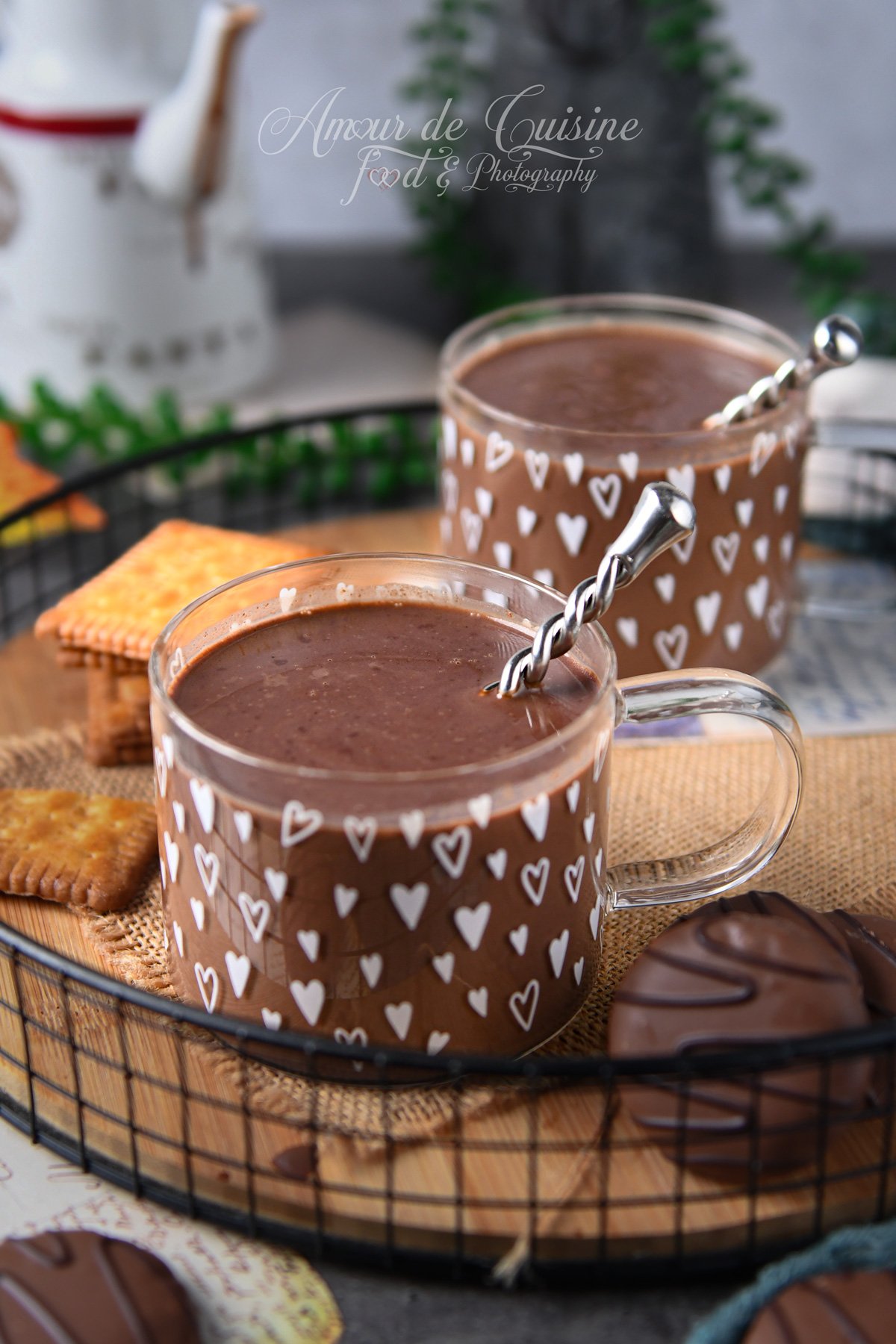Deux tasses en verre décorées de cœurs, remplies de chocolat chaud parisien épais, avec une cuillère torsadée, posées sur un plateau avec biscuits et douceurs au chocolat.