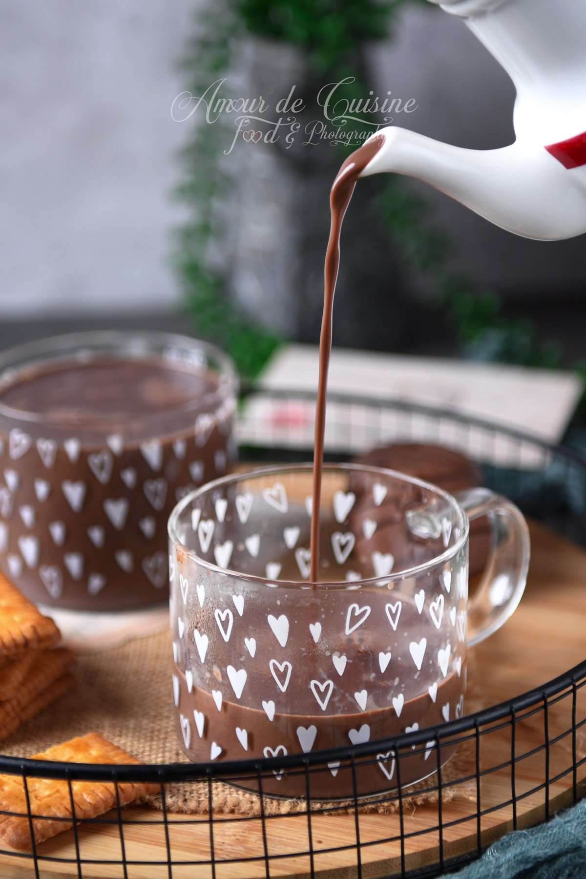 Un chocolat chaud parisien épais est versé d’une petite théière dans une tasse en verre décorée de cœurs, sur un plateau avec biscuits, avec une seconde tasse en arrière-plan.
