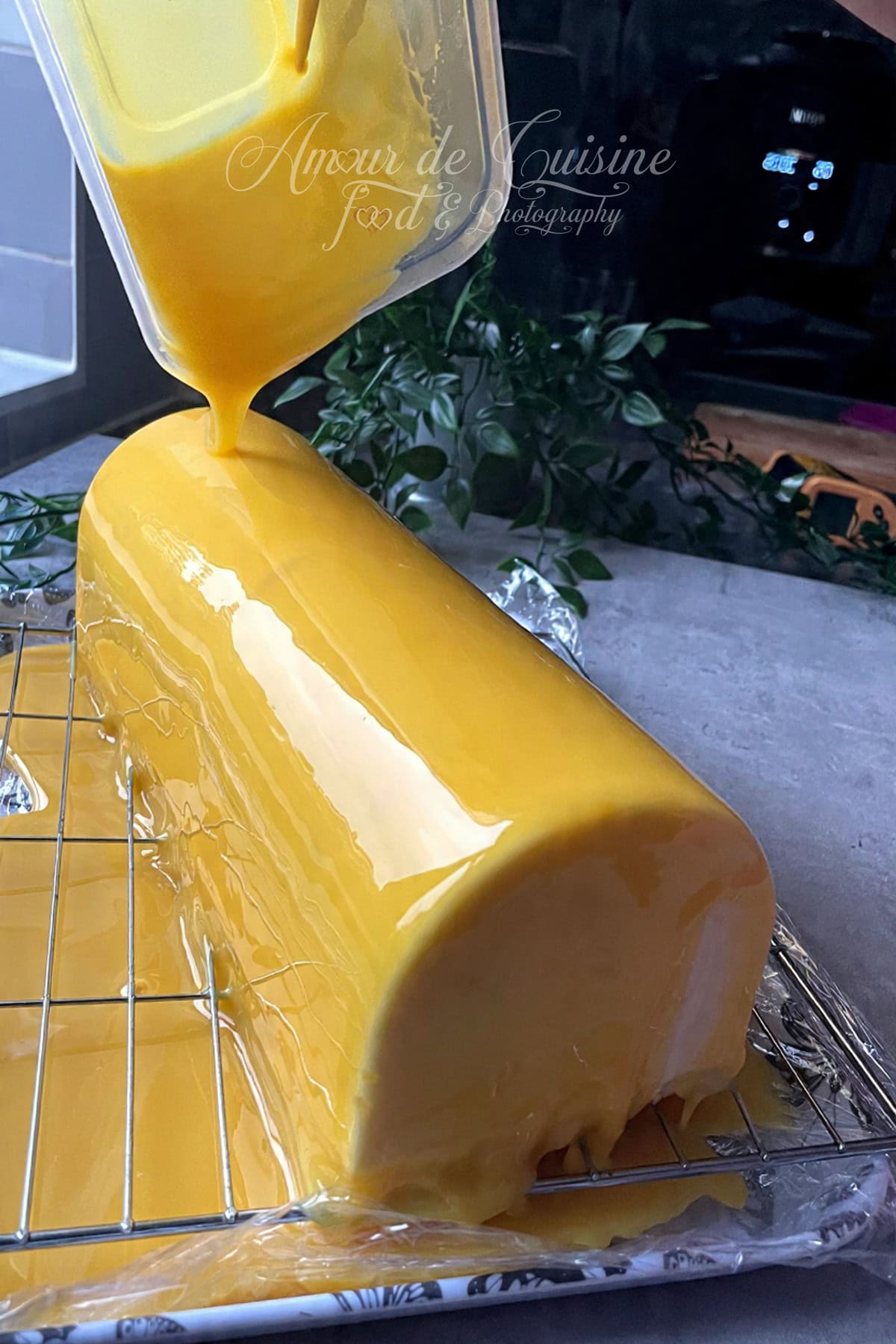 Glaçage miroir jaune versé sur une bûche mangue coco bien congelée, posée sur une grille au-dessus d’un plateau, pour obtenir une finition lisse, brillante et régulière.