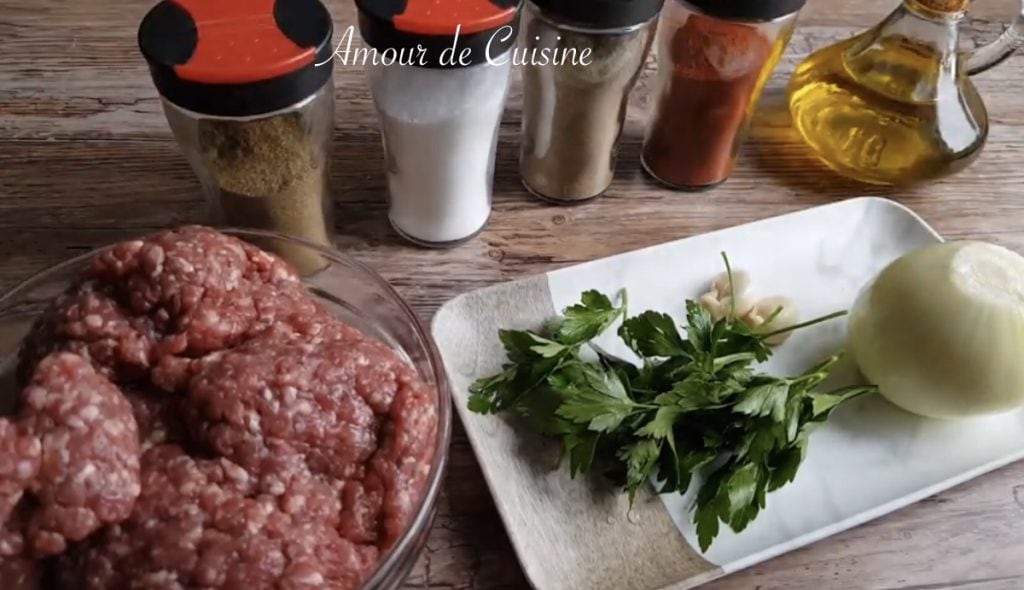Bol en verre rempli de viande hachée crue posé sur une table en bois, à côté d’un plateau avec un oignon entier, gousses d’ail et bouquet de persil frais, avec au fond plusieurs pots d’épices alignés et une carafe d’huile d’olive.
