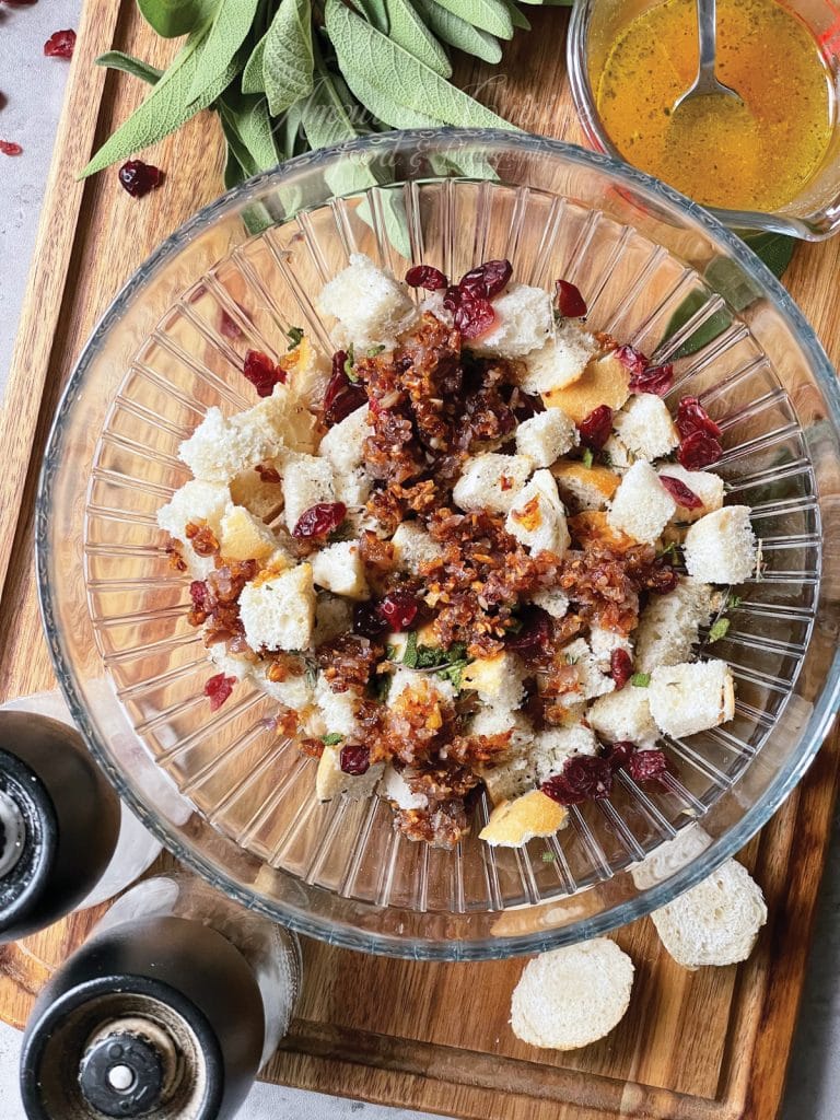 Grand saladier en verre rempli de cubes de pain blanc, canneberges séchées et oignons revenus, posé sur une planche en bois avec feuilles de sauge fraîche, petit pichet de beurre fondu et moulins à sel et poivre autour.
