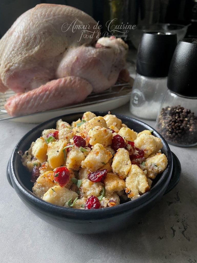 Bol noir rempli de farce à la baguette croustillante, canneberges rouges et herbes fraîches posé sur un plan de travail, avec une dinde crue prête à rôtir sur grille au second plan et moulins à sel et poivre flous.