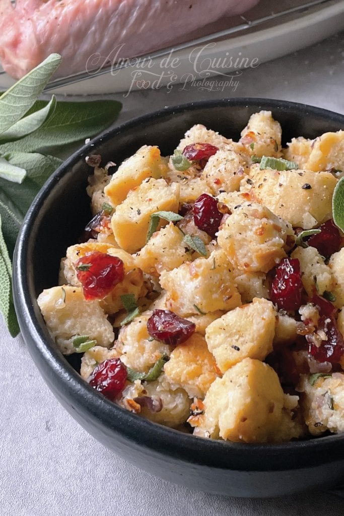 Gros plan sur un bol noir rempli de farce à base de cubes de pain dorés, canneberges rouges et petites feuilles de sauge, posé sur un plan de travail clair avec en arrière-plan une dinde crue floue sur sa grille.