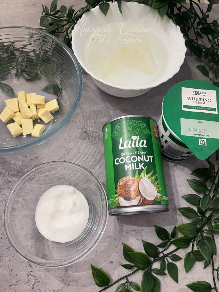 Ingrédients du crémeux coco sur plan de travail : morceaux de chocolat blanc, sucre, lait de coco en conserve, crème liquide et gélatine hydratée dans un bol, prêts pour la recette.