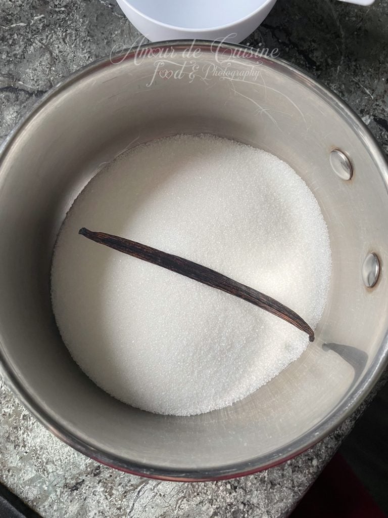 Casserole en inox vue de dessus remplie de sucre blanc en poudre avec une gousse de vanille entière posée au centre, sur un plan de travail gris, bol blanc flou en arrière-plan.