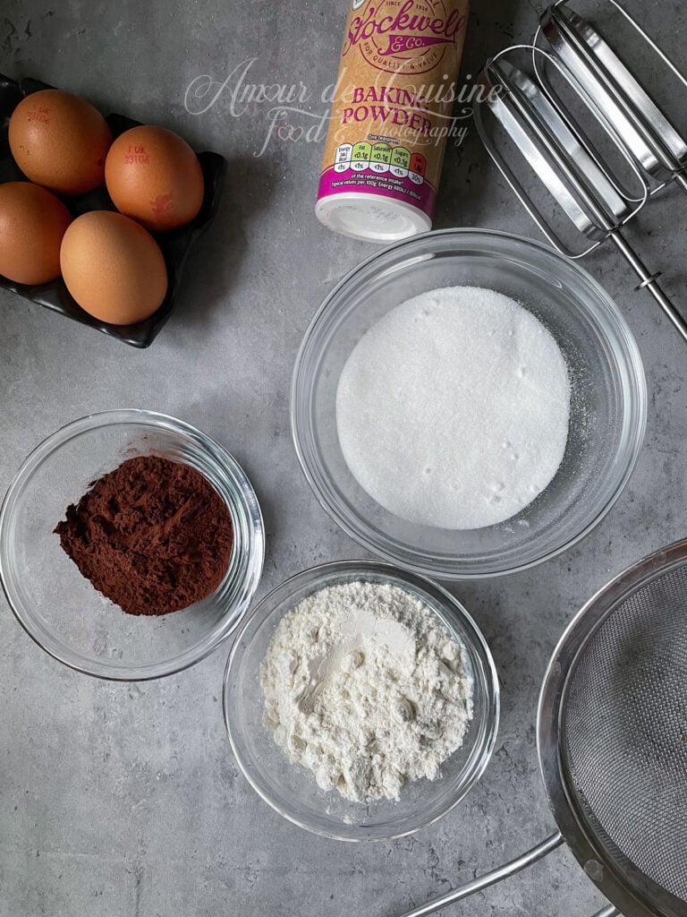 Ingrédients pour la génoise cacao d’une bûche de Noël : bols de sucre, farine et cacao en poudre, œufs entiers, levure chimique et fouets électriques posés sur un plan de travail gris avec un tamis en métal.