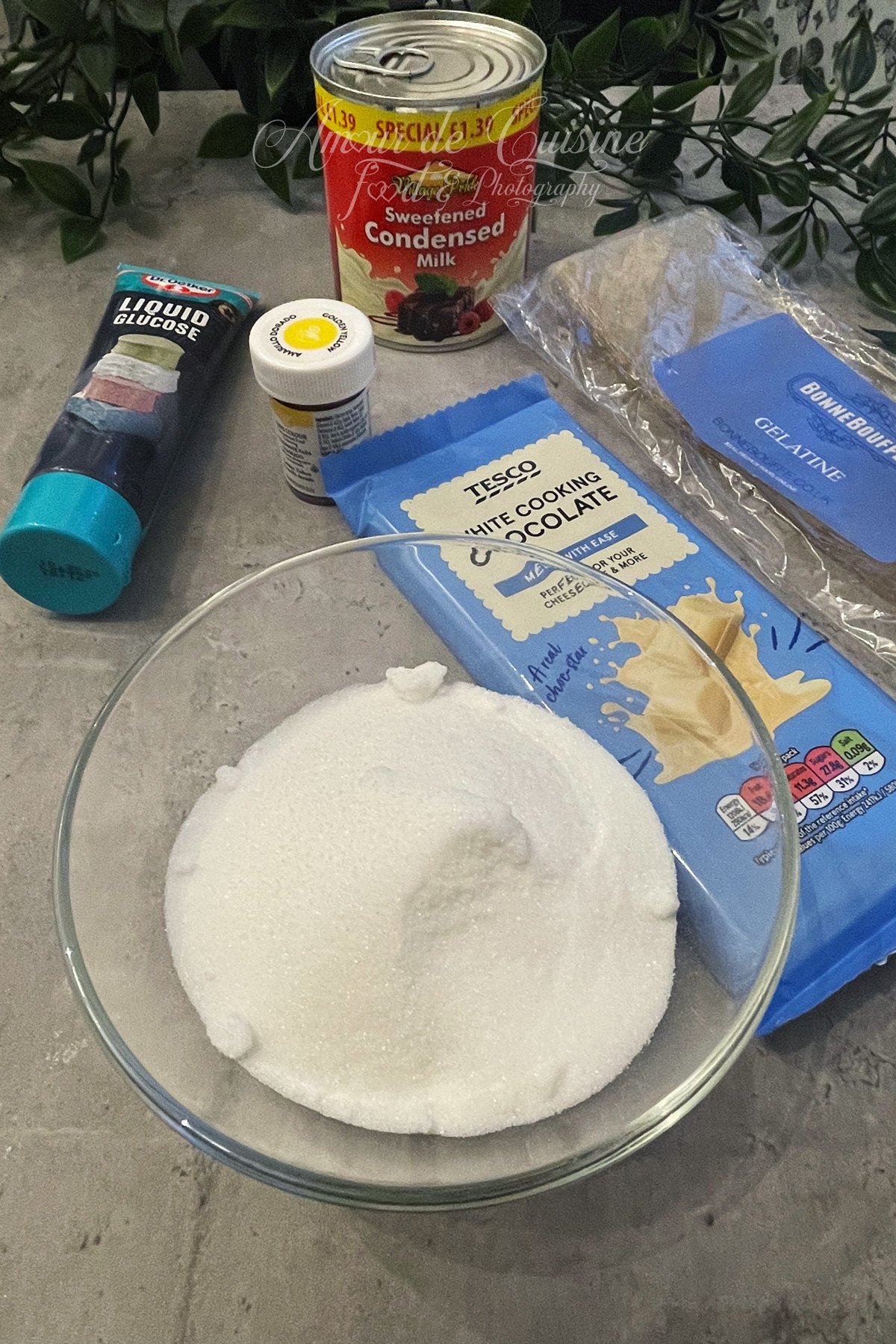 Ingrédients du glaçage miroir sur le plan de travail : sucre, glucose liquide, chocolat blanc, lait concentré sucré, gélatine, colorant jaune et eau, prêts pour réaliser un glaçage brillant.
