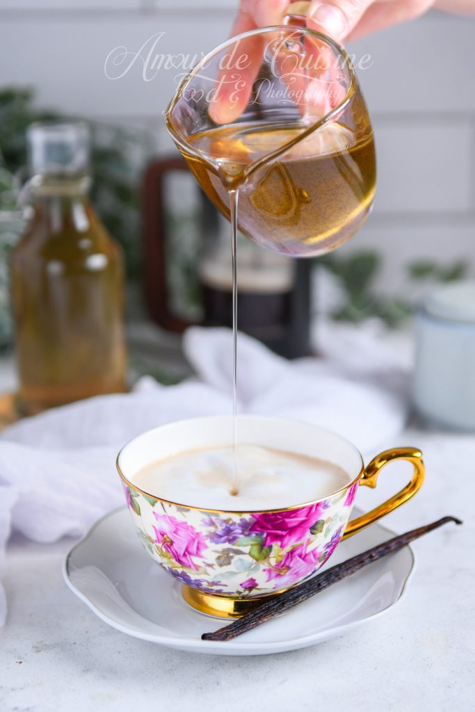 Main tenant un petit pichet en verre versant un filet de sirop de vanille maison dans un latte mousseux servi dans une tasse à fleurs avec anse dorée, posée sur une soucoupe avec une gousse de vanille.
