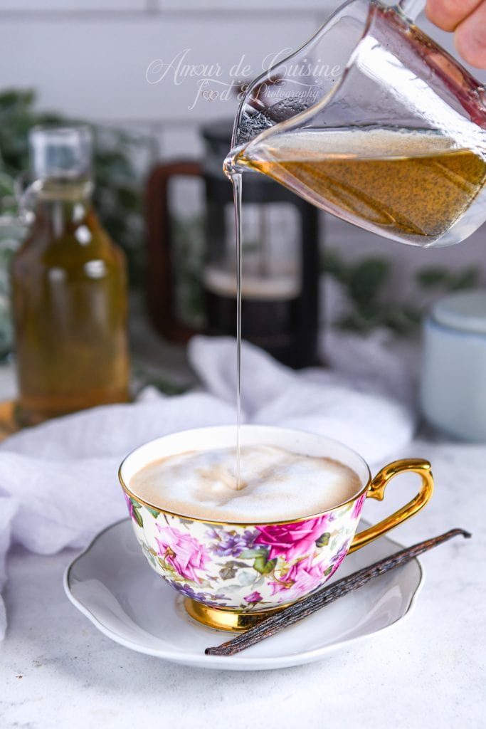 Pichet en verre versant un filet de sirop de vanille maison dans une tasse de latte mousseux à fleurs et anse dorée, posée sur une soucoupe avec une gousse de vanille, arrière-plan de cuisine flou.