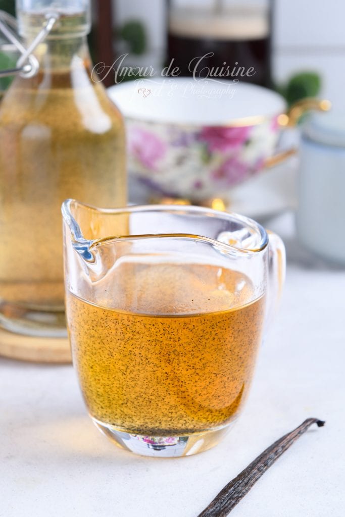 Petit pichet en verre rempli de sirop de vanille maison doré aux grains visibles, posé sur un plan de travail clair avec une gousse de vanille au premier plan et une bouteille et tasse floues en arrière-plan.