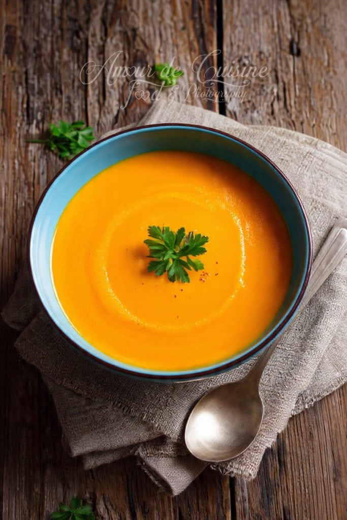 Bol de soupe de légumes rôtis, lisse et orangée, garni d’un brin de persil frais, posé sur un linge en lin sur une table en bois rustique.