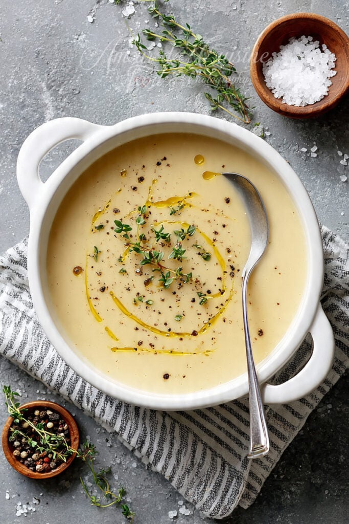 Vue du dessus d’une soupe de pommes de terre veloutée dans un bol blanc, avec un filet d’huile d’olive, du thym frais et du poivre, posée sur un torchon rayé avec une cuillère.