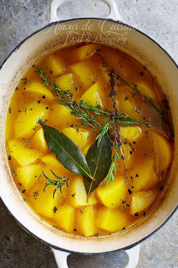 Cocotte en fonte avec des morceaux de pommes de terre qui mijotent dans un bouillon doré, avec feuille de laurier, brins de thym et romarin.