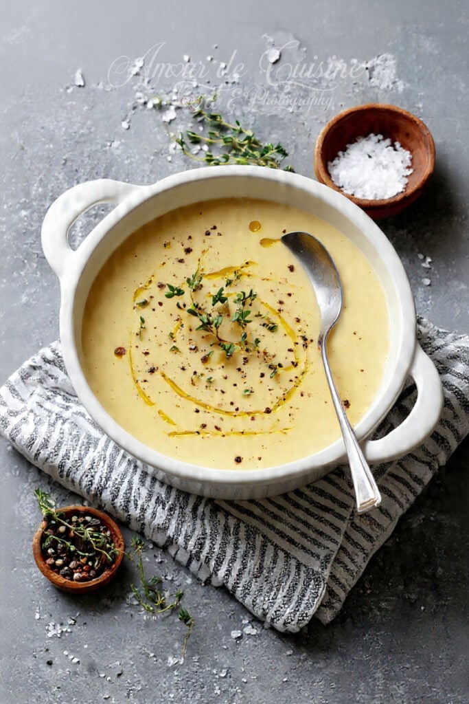 Bol de soupe de pommes de terre veloutée dans un plat blanc, garni de thym, de poivre et d’un filet d’huile d’olive, avec une cuillère, du sel en flocons et un petit bol d’épices sur une table grise.