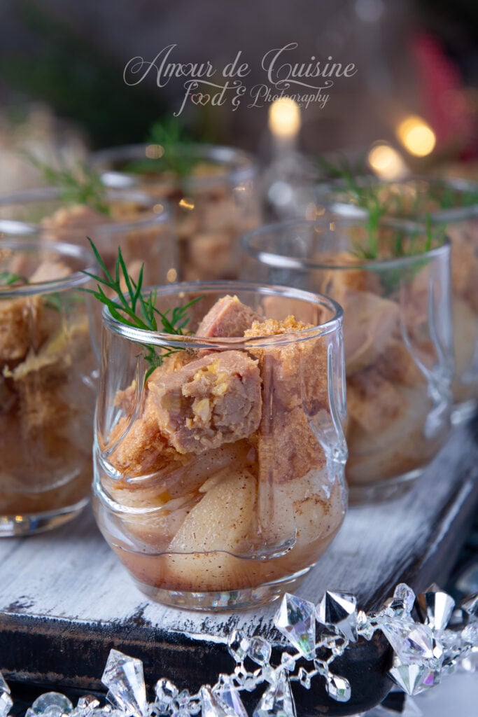 Gros plan sur une verrine de foie gras, poires épicées et miettes de pain d’épices, décorée d’un brin d’aneth, posée sur un plateau en bois avec une guirlande de cristaux scintillants.