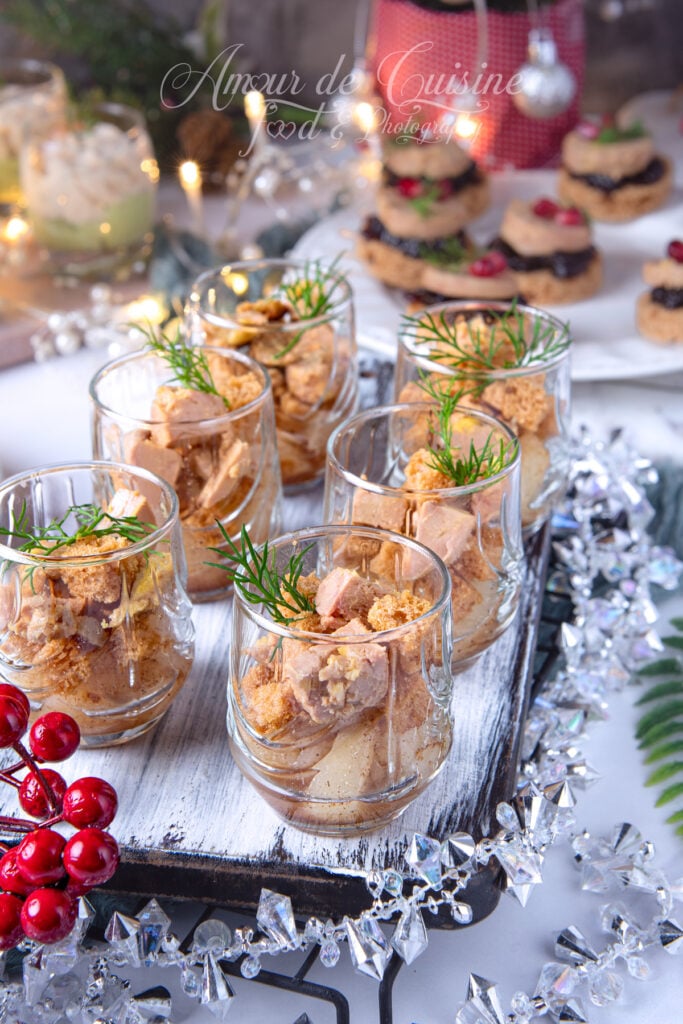 Verrines de foie gras, poire et pain d’épices servies dans de petits verres sur un plateau en bois, décor de Noël scintillant en arrière-plan avec guirlandes et lumières floues.
