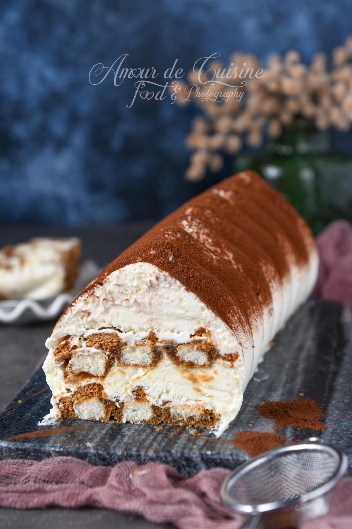 Bûche tiramisu au mascarpone, roulée et saupoudrée de cacao, avec une tranche coupée montrant les couches de crème et biscuits cuillère imbibés, posée sur une ardoise.