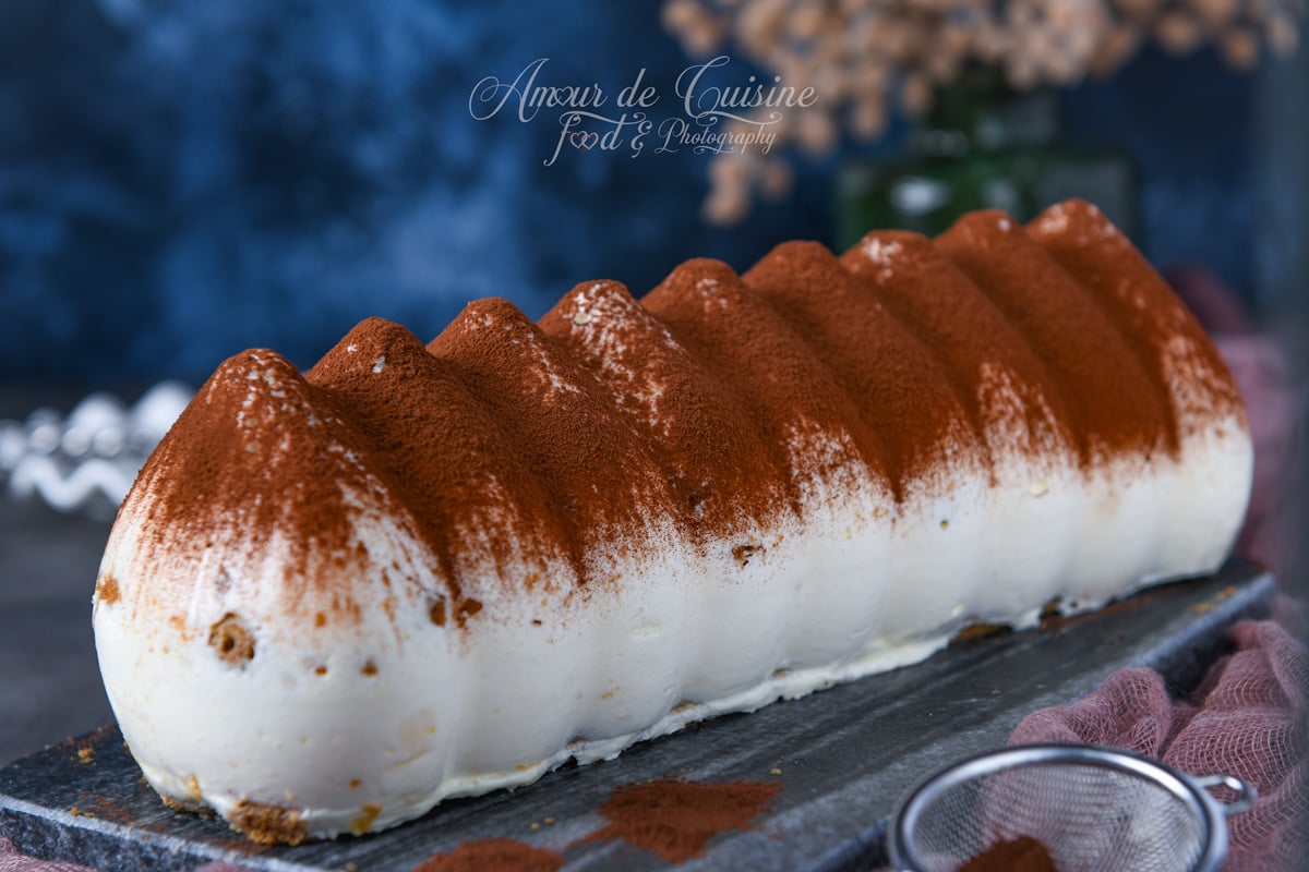 Bûche tiramisu au mascarpone, moulée en relief et généreusement saupoudrée de cacao, présentée entière sur une planche en ardoise, avec un tamis et un voile rose en décor.