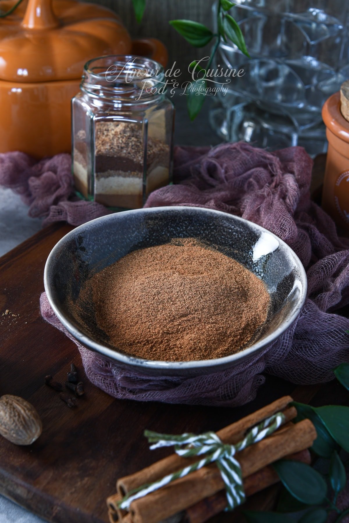 Bowl of homemade pumpkin pie spice on a wooden board, styled with cinnamon sticks tied in twine, whole nutmeg and cloves, a mauve cloth, and a glass jar of spice blend in a warm, autumn kitchen setting.
