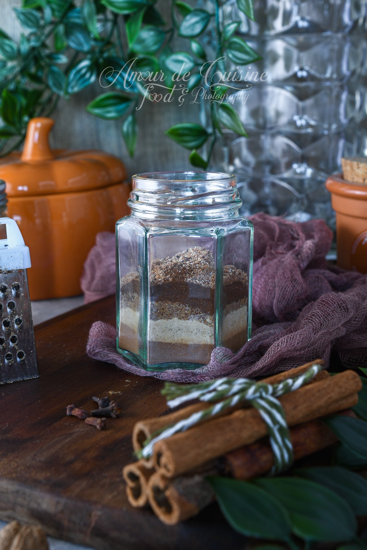 Homemade pumpkin pie spice layered in a clear glass jar on a wooden board, with cinnamon sticks tied in twine, whole cloves, a small grater, and a rustic kitchen background with green leaves and a mauve cloth.