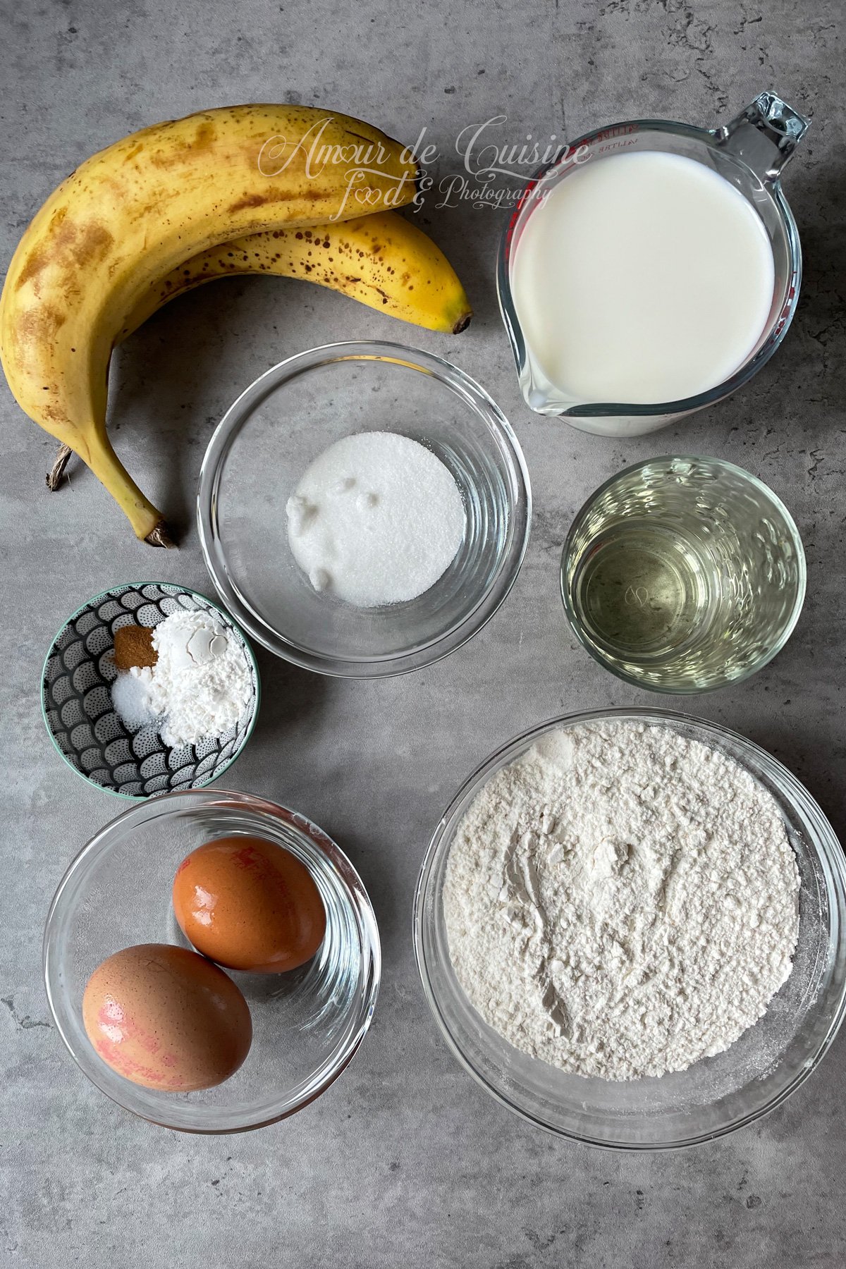 Banana pancake ingredients arranged on a countertop: two ripe bananas, milk in a measuring jug, oil, flour, sugar, two eggs, and a small bowl with baking powder, salt, and cinnamon.