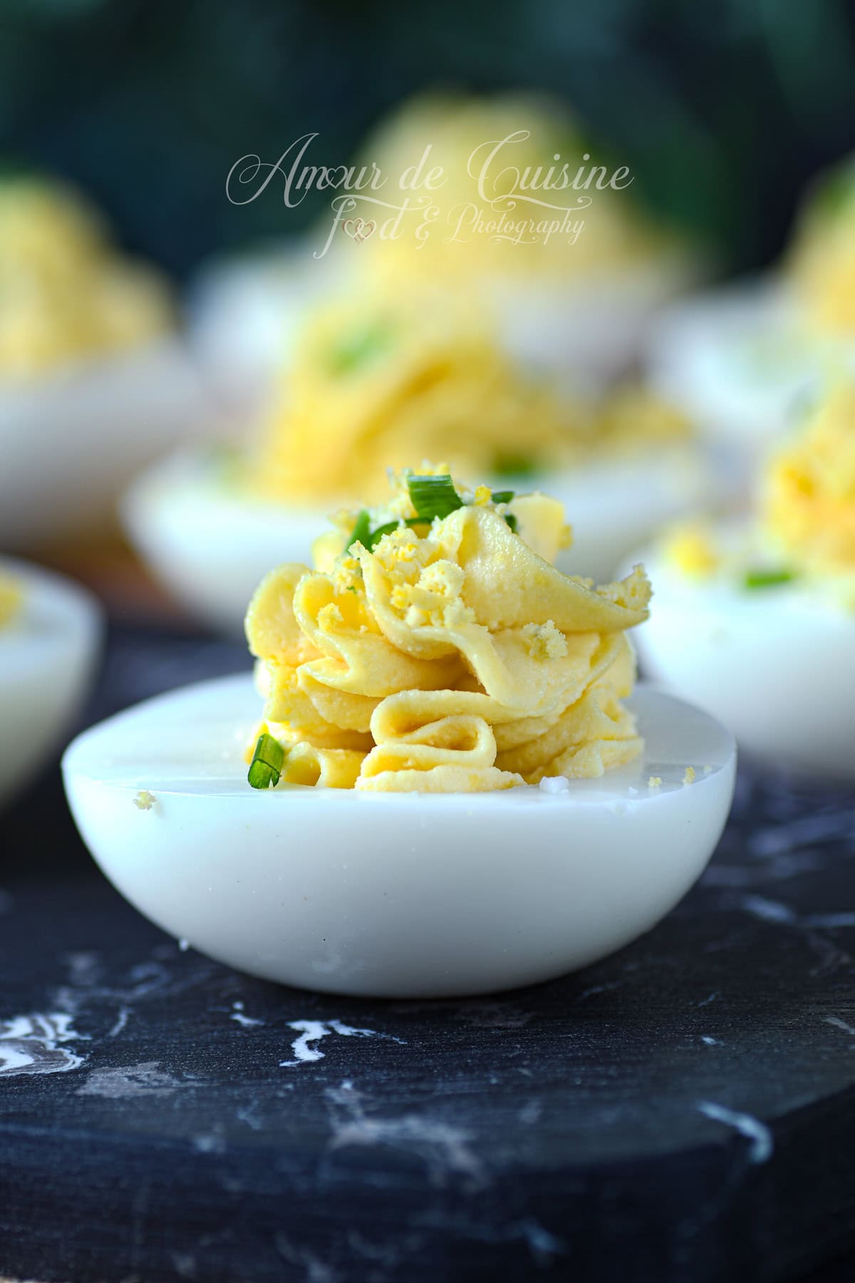 Gros plan sur un œuf mimosa : demi-oeuf dur garni d’une crème de jaune pochée en rosace, parsemé de jaune émietté et de ciboulette, sur une planche sombre effet marbre.